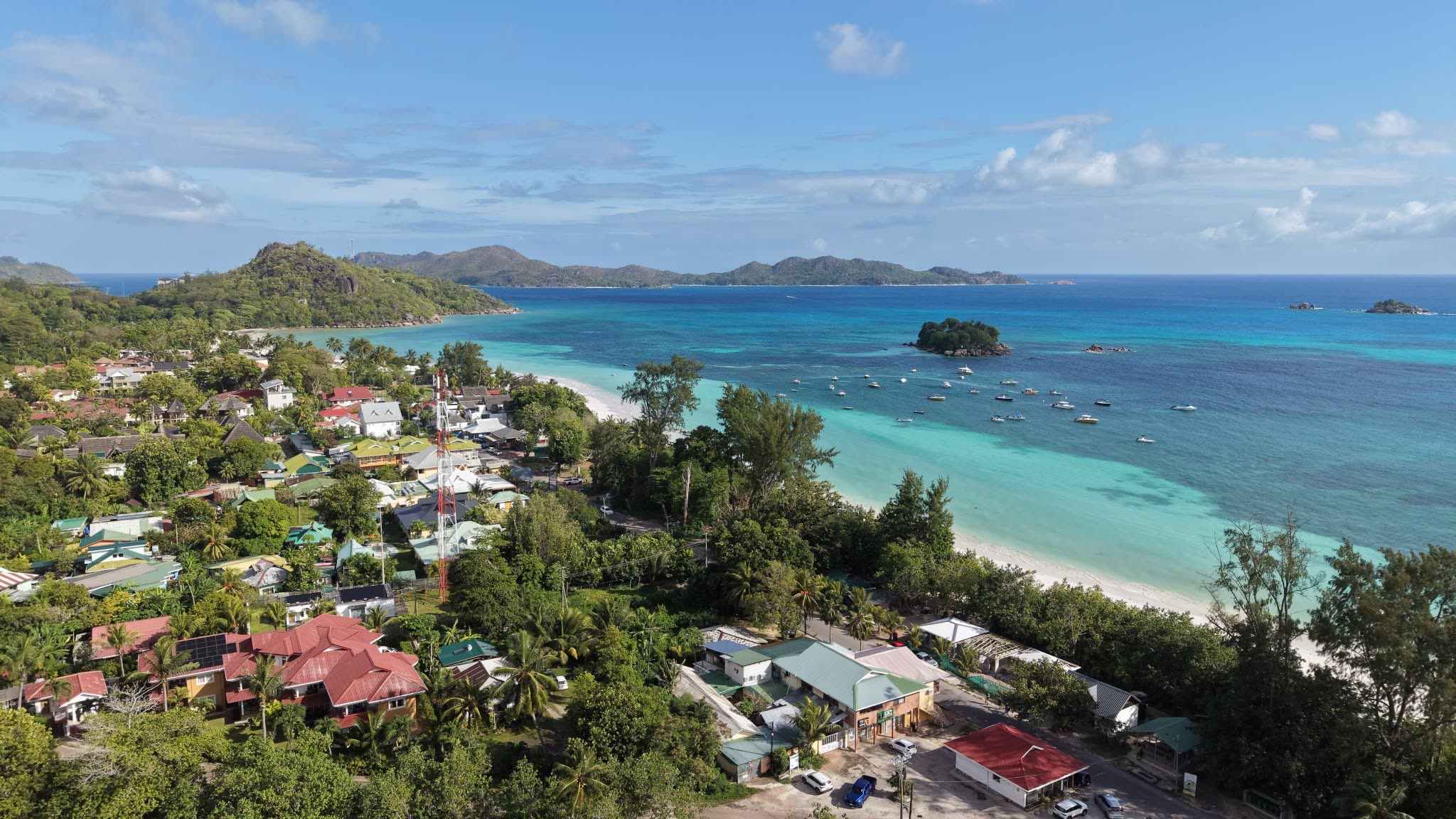 Aerial view of a village, a long white-sand beach and a turquoise bay with boats at anchor and wooded hills beyond