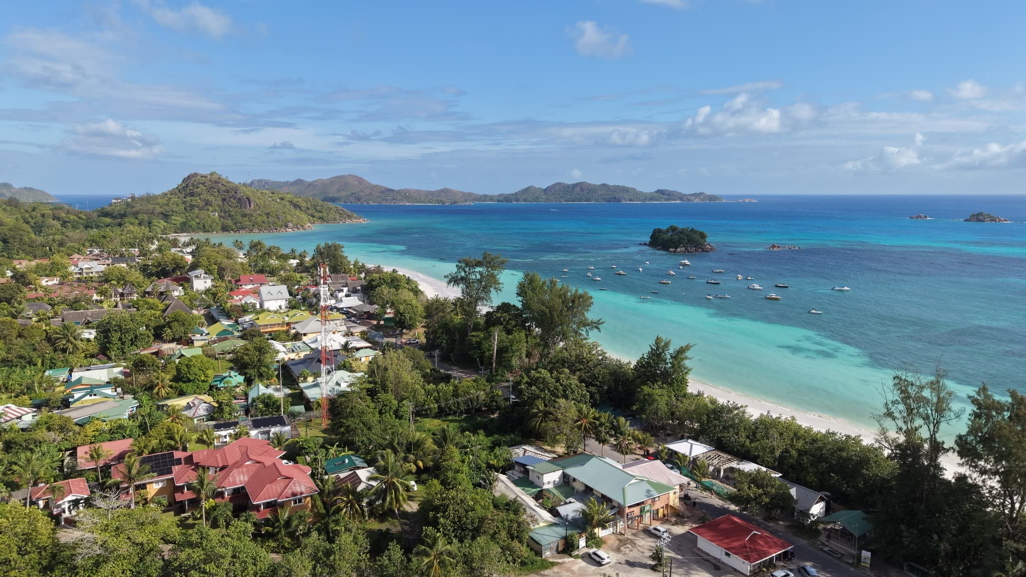 Aerial view of a village and a red-and-white mast beside a long white-sand beach, with a turquoise bay, boats at anchor and islands beyond