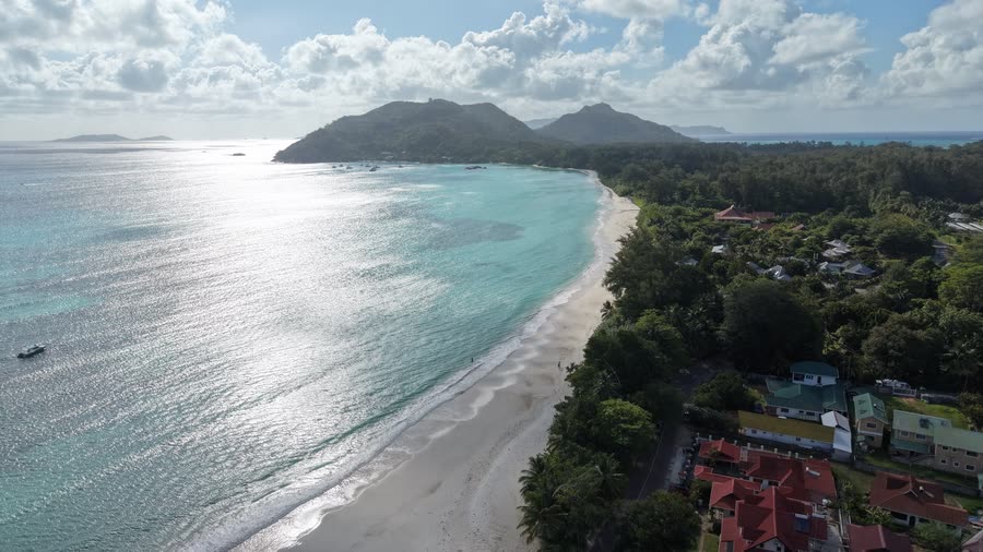 Aerial view of a long white-sand beach curving along turquoise water, with wooded hills at its far end and red-roofed houses among the trees beside it